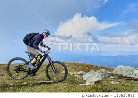 Cyclist man riding electric mountain bike outdoors. Male tourist biking along grassy trail in the mountains, wearing helmet and backpack. Concept of sport, active leisure and nature. Cyclist man riding electric mountain bike outdoors. Male tourist biking along grassy trail in the mountains, wearing helmet and backpack. Concept of sport, active leisure and nature. 113623931