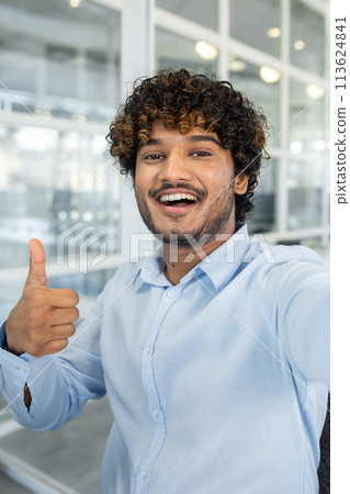 A cheerful young man with curly hair gives a thumbs up in a modern office setting, exuding confidence and positivity. Perfect for business and motivational themes. 113624841