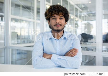 Portrait of a confident young man with curly hair, dressed in a light blue shirt, sitting in a well-lit office environment. Arms crossed, he exudes professionalism and approachability. Portrait of a confident young man with curly hair, dressed in a light blue shirt, sitting in a well-lit office environment. Arms crossed, he exudes professionalism and approachability. 113624848