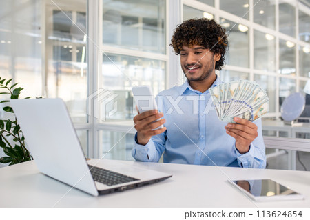 Joyful male entrepreneur in a modern office environment, holding a mobile phone and multiple US dollar bills, smiling and looking at phone. 113624854