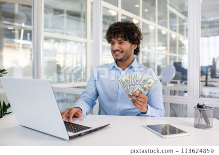 A cheerful young man in a blue shirt holds US dollar bills while sitting at a desk with a laptop in a bright office setting, showcasing financial success. A cheerful young man in a blue shirt holds US dollar bills while sitting at a desk with a laptop in a bright office setting, showcasing financial success. 113624859