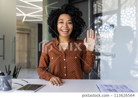 Cheerful young woman engaging in a video call, waving to the camera with a big smile, well-lit modern office background. Cheerful young woman engaging in a video call, waving to the camera with a big smile, well-lit modern office background. 113626564