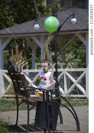 a boy with a green balloon stands near a bench in a city park 113626937