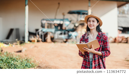 Optimistic young farmer holding a notepad stands in front of farm equipment, ready to tackle the day's tasks. 113634772
