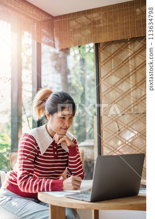 Pensive woman with a ponytail, sitting at a wooden table with a laptop, in a sunlit room with blinds. Pensive woman with a ponytail, sitting at a wooden table with a laptop, in a sunlit room with blinds. 113634798