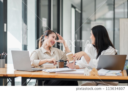 One woman laughs with her hand on her forehead while another hands her a coffee cup, both seated at a work desk with laptops. 113634807