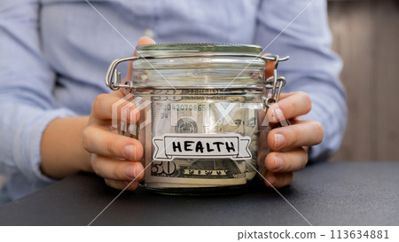 Female hands Saving Money In Glass Jar filled with Dollars banknotes. HEALTH transcription in front of jar. Managing personal finances extra income for future insecurity 113634881
