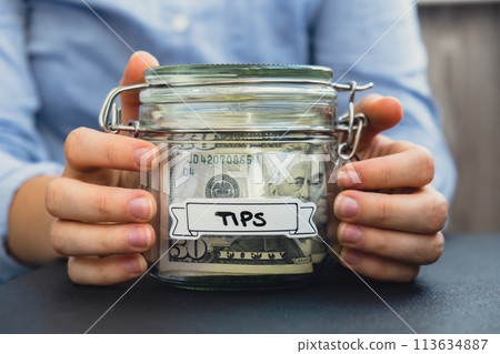 Female hands Saving Money In Glass Jar filled with Dollars banknotes. TIPS transcription in front of jar. Managing personal finances extra income for future insecurity 113634887