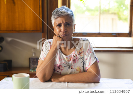 A mature pensive biracial woman sits at a home kitchen table on a video call 113634907