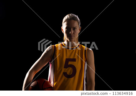 Young Caucasian female basketball player poses confidently in a basketball uniform on a black backgr 113634916