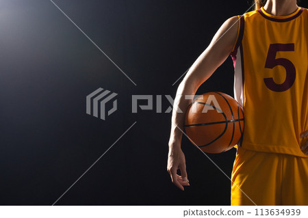 Athlete in basketball uniform holds a ball on a black background, with copy space 113634939