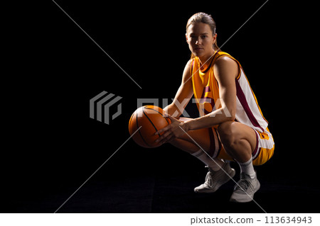 Young Caucasian female basketball player crouches in a basketball pose on a black background, with c 113634943