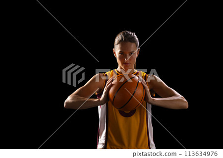 Young Caucasian female basketball player holds a basketball confidently on a black background 113634976