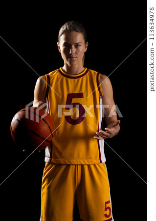 Confident young Caucasian female basketball player poses in a basketball uniform on a black backgrou 113634978