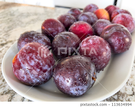 A plate of ripe, frosty plums sits prominently on a marble countertop in a sunlit kitchen with a crisp, white interior and neatly arranged appliances. A plate of ripe, frosty plums sits prominently on a marble countertop in a sunlit kitchen with a crisp, white interior and neatly arranged appliances. 113634994