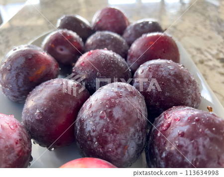 A plate of ripe, frosty plums sits prominently on a marble countertop in a sunlit kitchen with a crisp, white interior and neatly arranged appliances. A plate of ripe, frosty plums sits prominently on a marble countertop in a sunlit kitchen with a crisp, white interior and neatly arranged appliances. 113634998