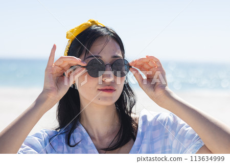 A young woman adjusts her sunglasses at the beach, unaltered A young woman adjusts her sunglasses at the beach, unaltered 113634999