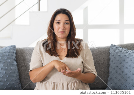 Young biracial woman gestures while sitting on a couch at home on a video call Young biracial woman gestures while sitting on a couch at home on a video call 113635101
