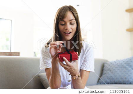 A young Caucasian woman appears pleasantly surprised as she opens a small red gift box 113635161