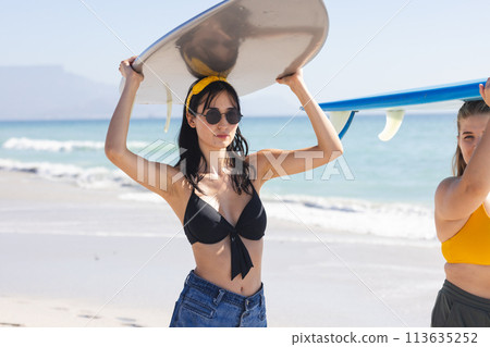 Young Caucasian woman and young biracial woman carry a surfboard on the beach 113635252