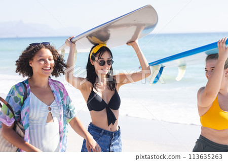 Young biracial and Caucasian women carry a surfboard on the beach Young biracial and Caucasian women carry a surfboard on the beach 113635263