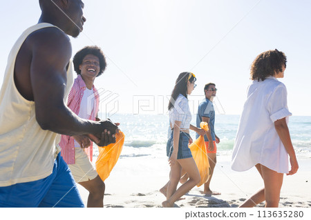 Diverse group of friends enjoy a day at the beach, collecting trash 113635280
