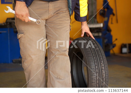 The mechanic technician is holding a new car tire to assemble it onto the wheel and the vehicle. The car mechanic is changing the car tire at the car repair shop or garage The mechanic technician is holding a new car tire to assemble it onto the wheel and the vehicle. The car mechanic is changing the car tire at the car repair shop or garage 113637144