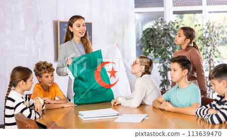 Positive young female teacher holding national flag of Algeria while working with preteens schoolgirls and schoolboys in classroom Positive young female teacher holding national flag of Algeria while working with preteens schoolgirls and schoolboys in classroom 113639470