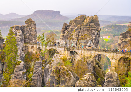 The Bastei Bridge stands tall among majestic sandstone rock formations with visitors enjoying the scenic view at sunset. Kurort Rathen, Saxon Switzerland, Germany The Bastei Bridge stands tall among majestic sandstone rock formations with visitors enjoying the scenic view at sunset. Kurort Rathen, Saxon Switzerland, Germany 113643903
