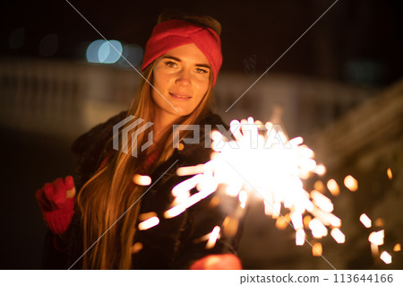 Woman holding sparkler night while celebrating Christmas outside. Dressed in a fur coat and a red headband. Blurred christmas decorations in the background. Selective focus 113644166