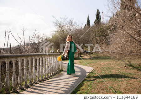 A woman in a green dress stands on a path next to a railing. She is holding a yellow bag and a yellow flower. The scene is peaceful and serene, with the woman posing for a photo. A woman in a green dress stands on a path next to a railing. She is holding a yellow bag and a yellow flower. The scene is peaceful and serene, with the woman posing for a photo. 113644169