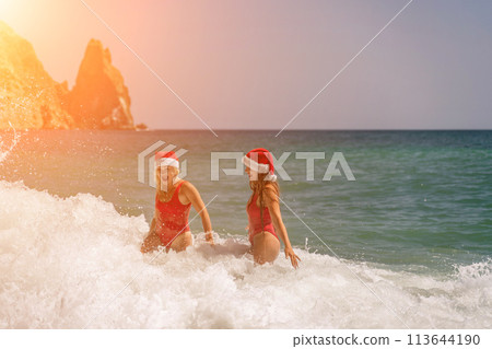 Women Santa hats ocean play. Seaside, beach daytime, enjoying beach fun. Two women in red swimsuits and Santa hats are enjoying themselves in the ocean waves. 113644190