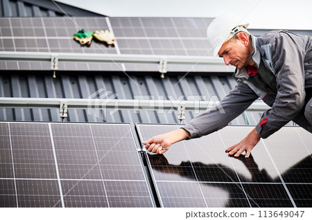 Man technician mounting photovoltaic solar panels on roof of house. Engineer in helmet installing solar module system with help of hex key. Concept of alternative, renewable energy. Man technician mounting photovoltaic solar panels on roof of house. Engineer in helmet installing solar module system with help of hex key. Concept of alternative, renewable energy. 113649047
