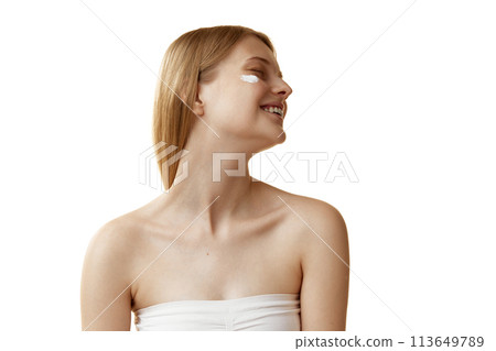 Young, blonde woman with cream swatches on cheeks looking away with smile enjoys self-care process against white studio background. 113649789