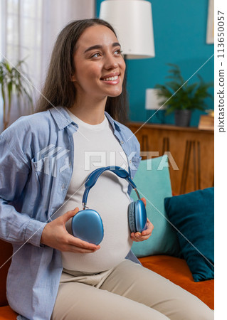 Pregnant woman sitting on home sofa listening music putting headphones on her tummy with child baby 113650057