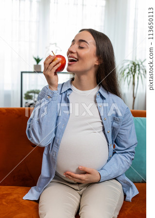 Happy pregnant woman eating fresh red apple fruit sitting on sofa healthy vitamin food motherhood 113650113