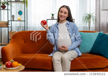 Happy pregnant woman eating fresh red apple fruit sitting on sofa healthy vitamin food motherhood 113650122