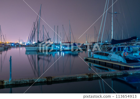 Night view of a boat dock with calm water and many boats and distant city lights in background Night view of a boat dock with calm water and many boats and distant city lights in background 113654913