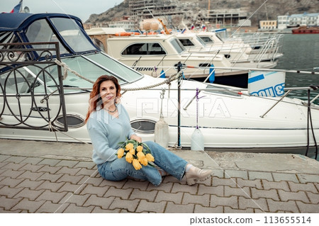 A woman sits on the dock with a bouquet of yellow flowers. The scene is peaceful and serene, with the woman looking out at the water. 113655514
