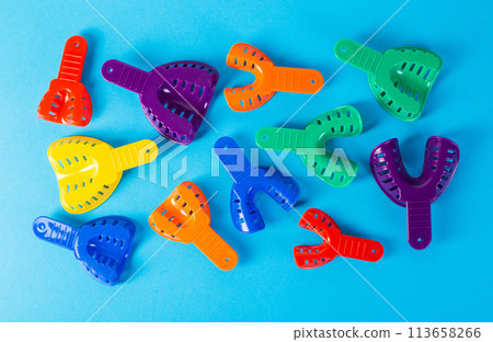 Multi-colored dental spoons for taking an impression of the dental jaw on a blue background. Orthodontics in dentistry. Dental prosthetics and crown manufacturing, close-up Multi-colored dental spoons for taking an impression of the dental jaw on a blue background. Orthodontics in dentistry. Dental prosthetics and crown manufacturing, close-up 113658266