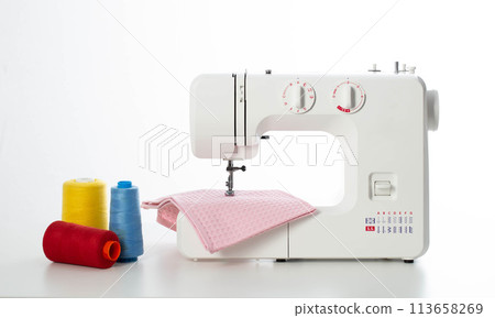 Modern automatic white sewing machine with pink fabric and multi-colored spools of thread on the table. White background. Tailoring and clothing repair services. Copy space for text Modern automatic white sewing machine with pink fabric and multi-colored spools of thread on the table. White background. Tailoring and clothing repair services. Copy space for text 113658269