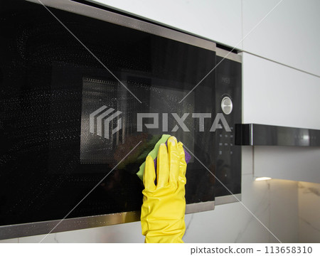 A girl's hand in a yellow glove with a sponge washes the microwave oven from grease and dirt. Foam and anti-grease. Cleaning household appliances in the kitchen 113658310
