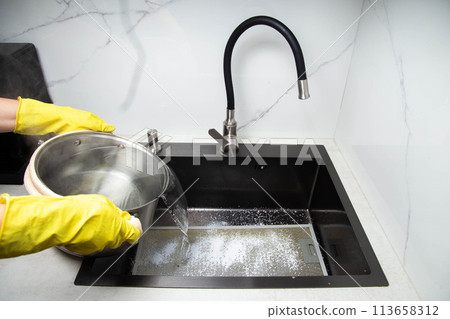 Man's hands in yellow gloves pour pots of hot water into a sink with dirty filters from the hood in the kitchen. Cleaning from dirt and grease with an oxygen cleaner. Copy space for text Man's hands in yellow gloves pour pots of hot water into a sink with dirty filters from the hood in the kitchen. Cleaning from dirt and grease with an oxygen cleaner. Copy space for text 113658312