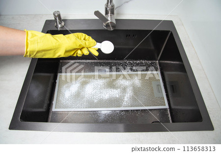 A man's hand in a yellow glove holds a spoon with oxygen bleach against the background of a kitchen sink. Cleaning from grease and dirt. Cleaning filters from kitchen hoods. 113658313