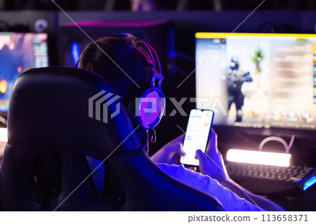Cybersport tournament Gamer wearing a headset plays video games on a PC in a computer club and holds a smartphone in his hand, multiplayer online combat competition in real-time strategy. Skill. Cybersport tournament Gamer wearing a headset plays video games on a PC in a computer club and holds a smartphone in his hand, multiplayer online combat competition in real-time strategy. Skill. 113658371