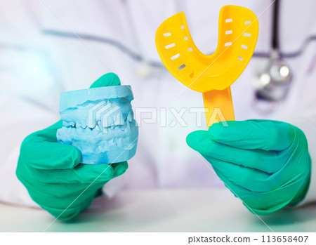Doctor dentist holds a yellow spoon and an impression of the patient's jaw in green medical gloves. The concept of making dentures and dental crowns based on a patient s impression in a dental 113658407