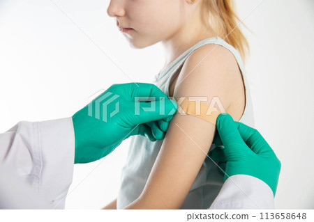 Doctor's hands in green medical gloves stick a plaster on the shoulder of a girl child after vaccination against hemophilus influenzae and pneumococcal. Copy space for text, revaccination Doctor's hands in green medical gloves stick a plaster on the shoulder of a girl child after vaccination against hemophilus influenzae and pneumococcal. Copy space for text, revaccination 113658648