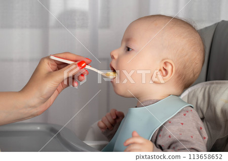 A mother feeds porridge from a spoon to a little boy who is 8 months old. Feeding infants. Copy space for text 113658652