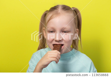 A beautiful joyful girl with white hair of seven years old eats a chocolate bar on a yellow background. Children are lovers of sweet junk food. Caries A beautiful joyful girl with white hair of seven years old eats a chocolate bar on a yellow background. Children are lovers of sweet junk food. Caries 113658668