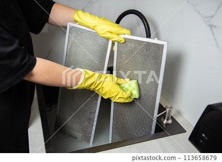 A girl's hands in yellow gloves with a sponge wash the kitchen hood filter with detergent to remove grease and dirt. Cleaning the kitchen, grease stains 113658679